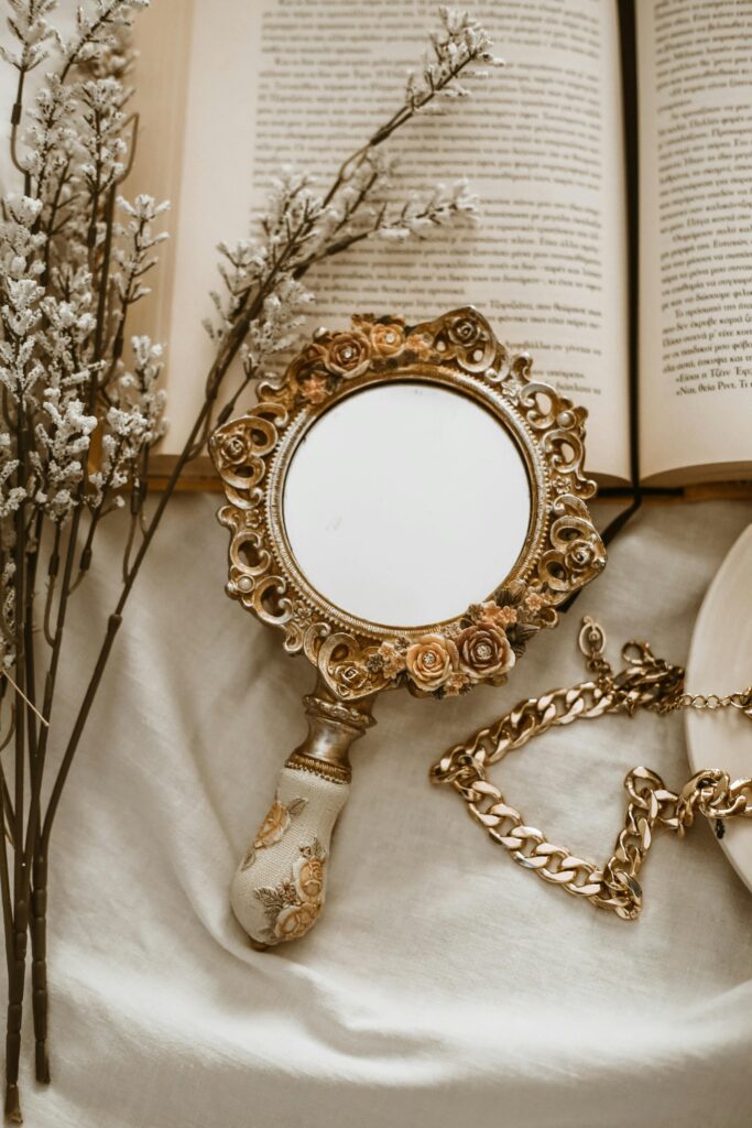 Vintage floral hand mirror beside books, gold jewelry, and delicate white flowers arranged on soft fabric.