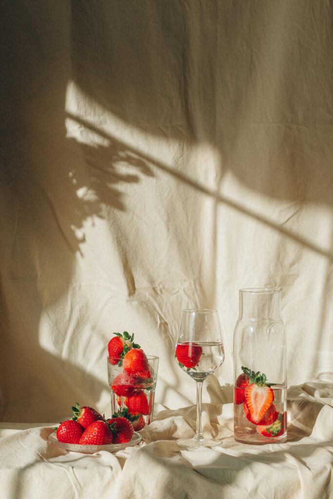 Fresh strawberries in glass cups and water bottles beside soft cream fabric with warm sunlight creating a cozy and romantic atmosphere.