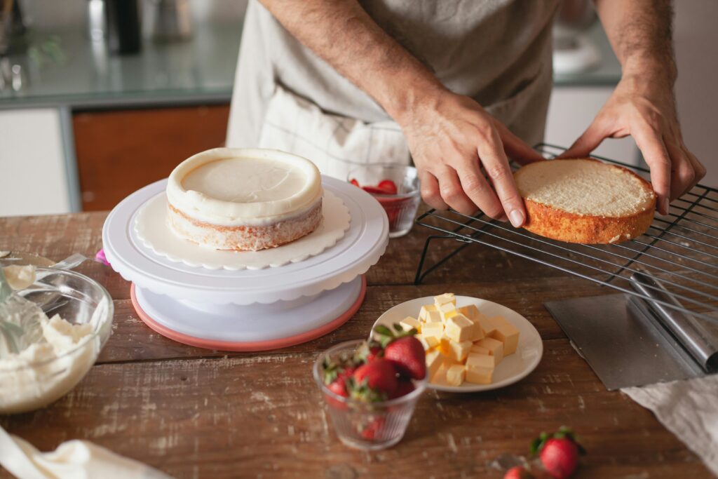 Person decorating a cake in a cozy kitchen with strawberries, butter cubes, and baking tools on a wooden table.