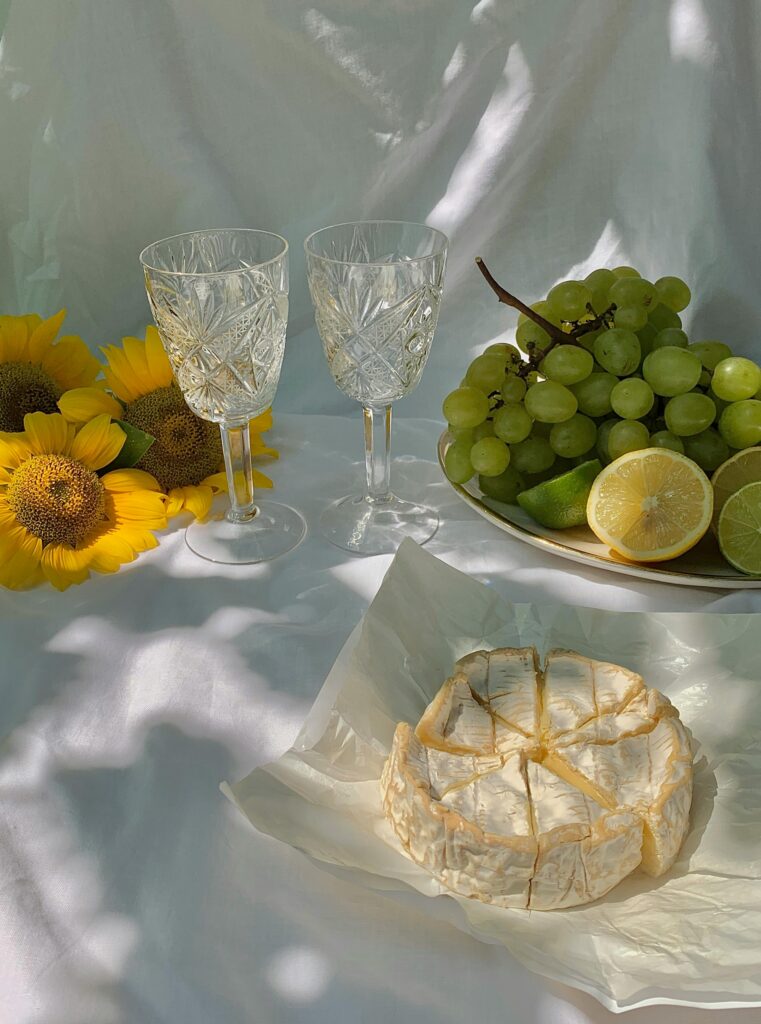 Pastry cake with grapes, glasses, and sunflowers arranged on a white picnic blanket with warm sunlight reflecting across the scene in a dreamy aesthetic setting.