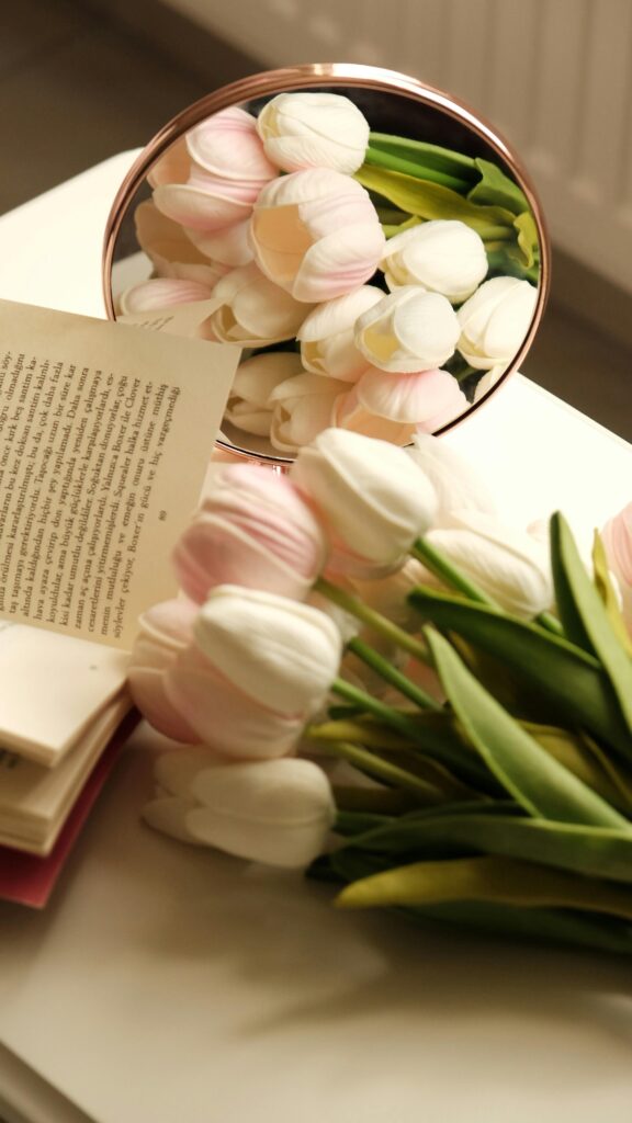 Pink tulips reflected in a mirror next to an open book in a soft cozy aesthetic setting with warm natural light.