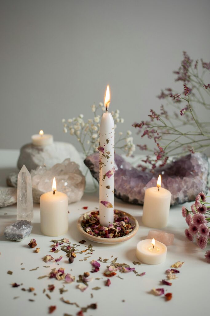 Three glowing candles surrounded by dried flower petals and manifestation stones in a warm cozy aesthetic setting for a calming self-care evening ritual.