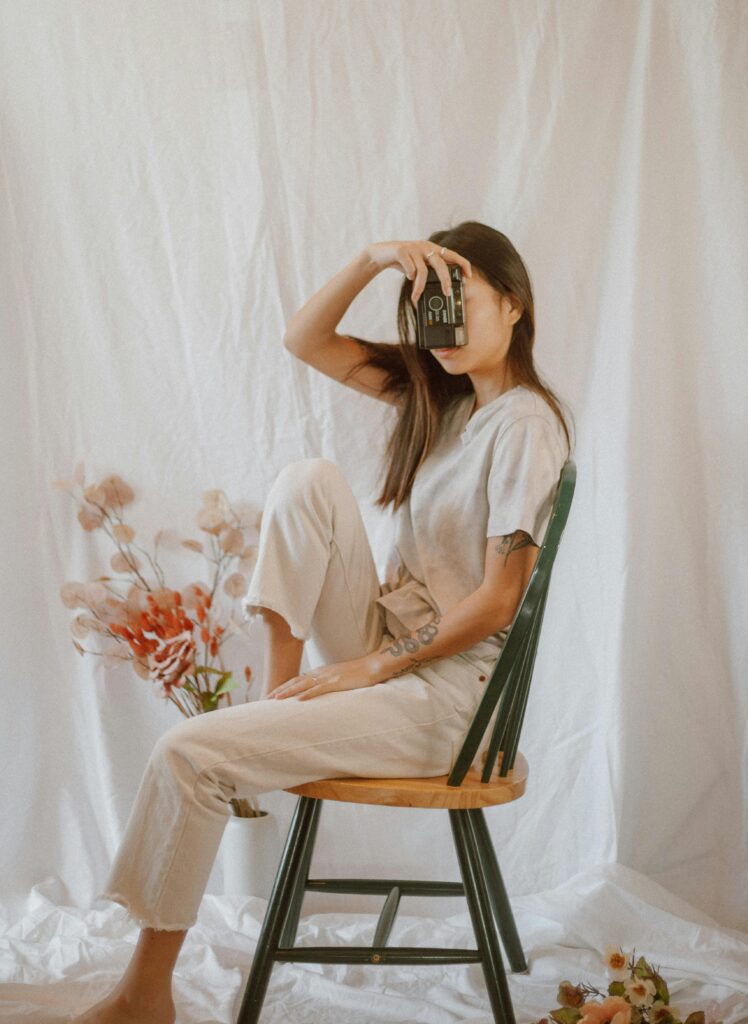 Girl wearing casual cozy clothes holding a vintage camera while surrounded by soft flowers in a dreamy warm aesthetic setting.