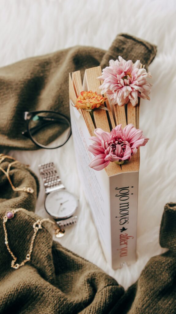 Jojo Moyes book with flowers used as a bookmark beside sunglasses and a wristwatch on a cozy aesthetic surface