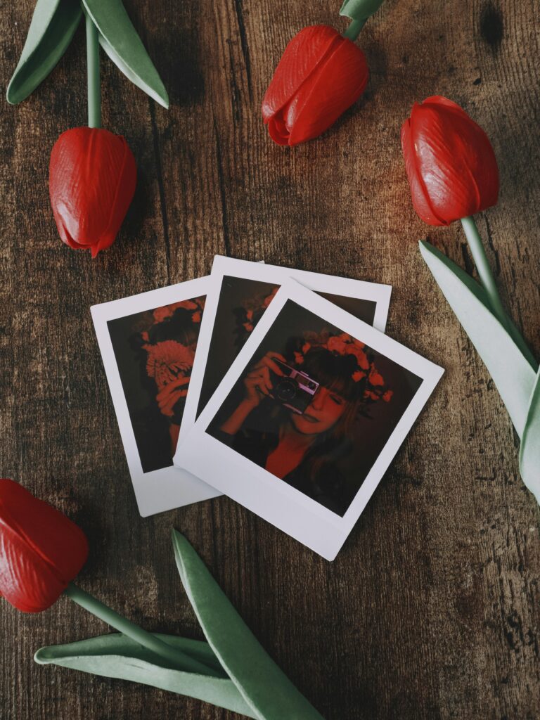 Red tulips surrounding vintage polaroid photos on a dark wooden table with cozy nostalgic aesthetic.