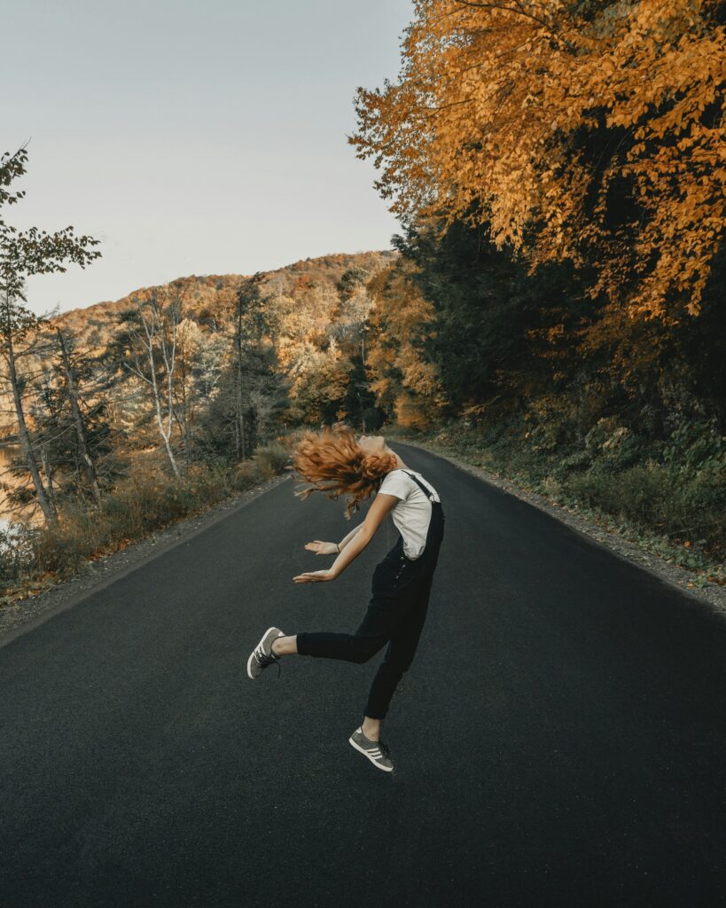 A woman dancing freely on an empty road during fall season surrounded by golden autumn leaves, warm sunlight, and a cozy cinematic atmosphere.