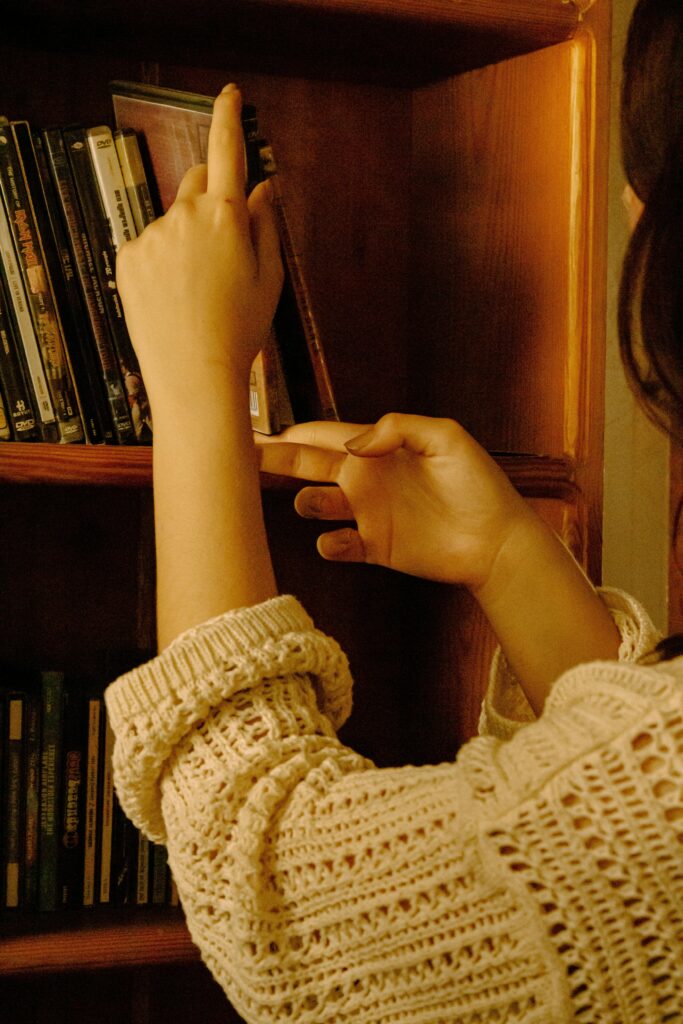 A cozy aesthetic image of a woman rearranging a bookshelf filled with books, candles, and small decor items in warm natural lighting, creating a peaceful slow living atmosphere.