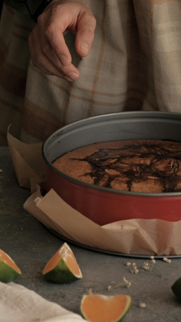 A cozy aesthetic photo of a freshly baked cake in a round baking pan with citrus slices on the counter and warm moody lighting.
