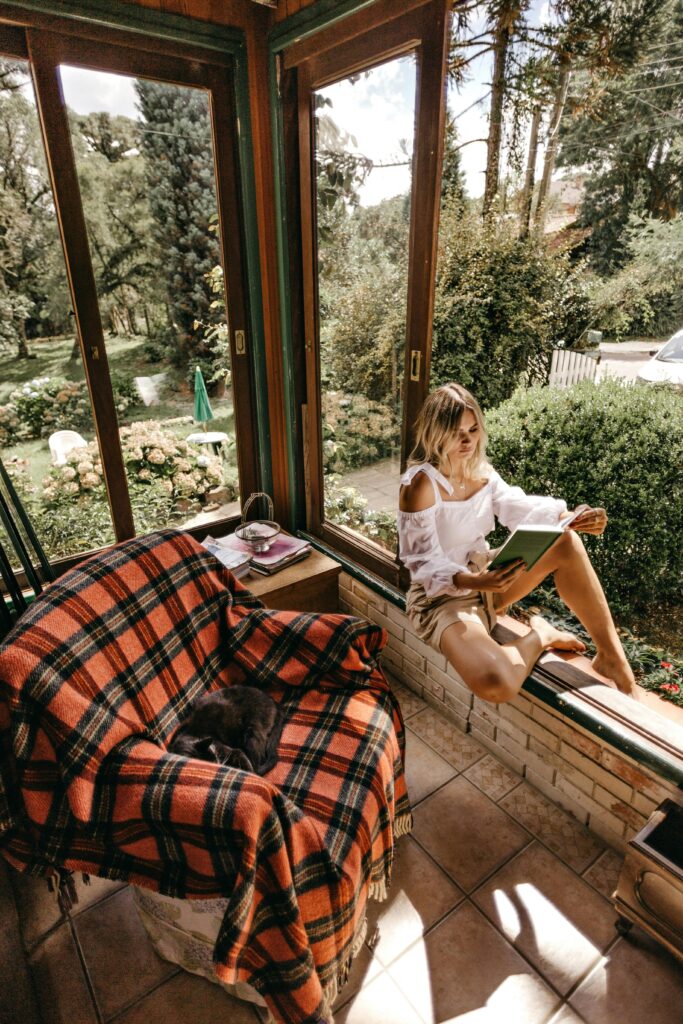Person reading a book beside a large sunlit window overlooking a peaceful garden.