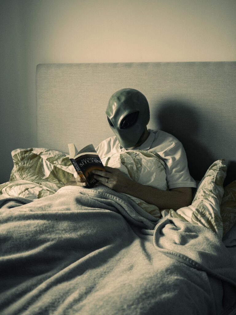 Person wearing an alien mask reading a book while sitting comfortably in bed under blankets.