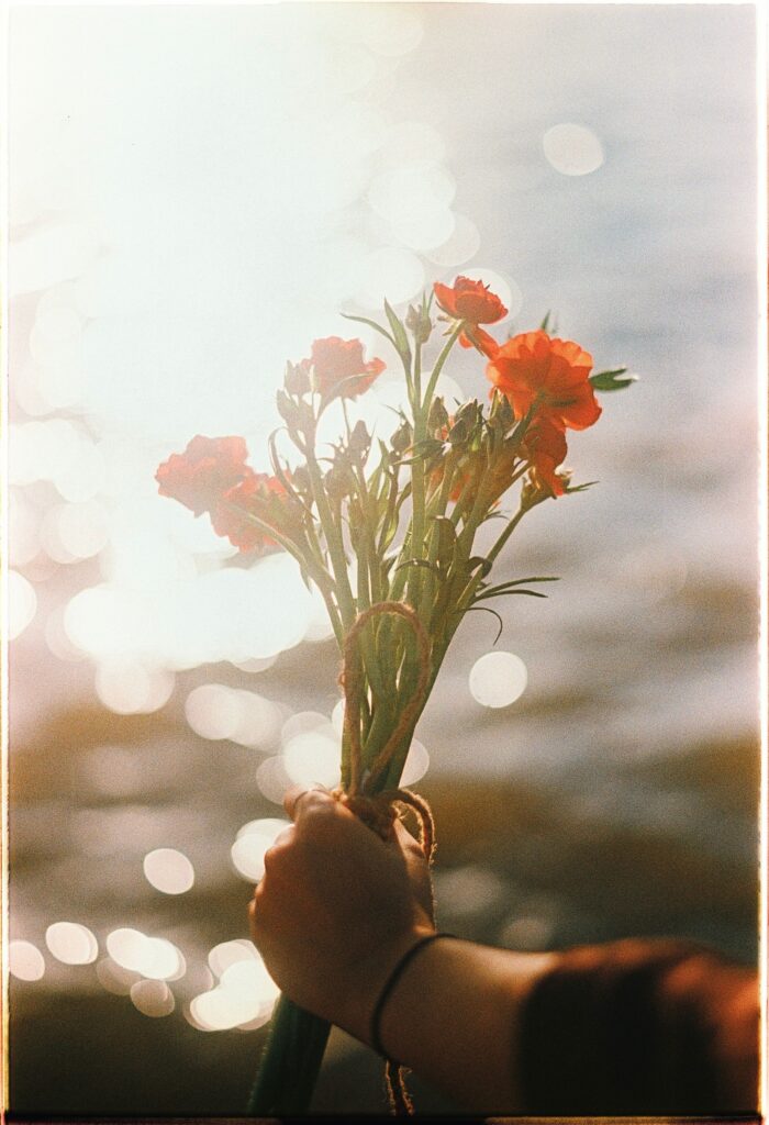 Hand holding a bouquet of orange flowers glowing in warm sunset light beside shimmering water.