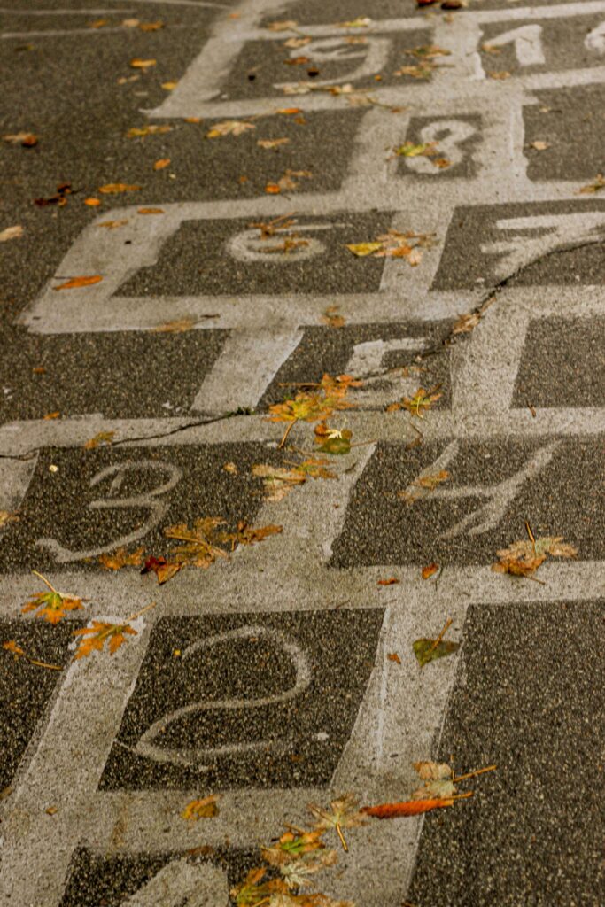 Weathered hopscotch grid drawn on pavement with scattered leaves, evoking nostalgic childhood summer memories.