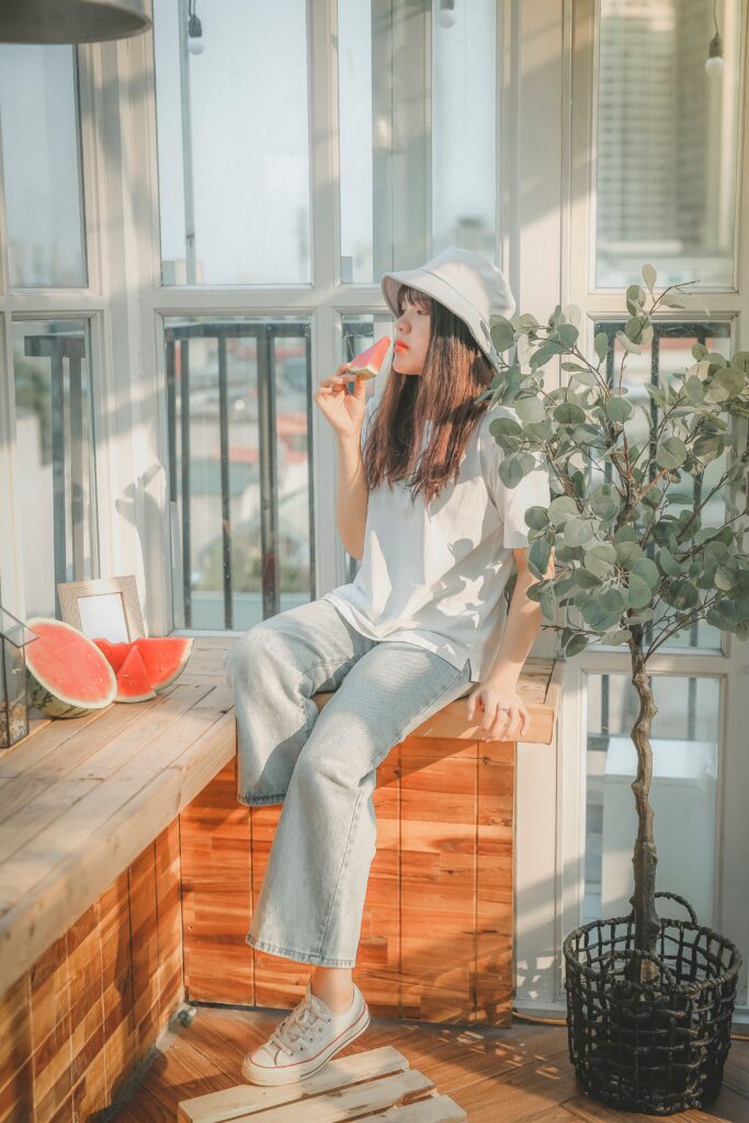 Young woman eating watermelon in soft afternoon sunlight beside large windows, creating a dreamy nostalgic summer atmosphere.