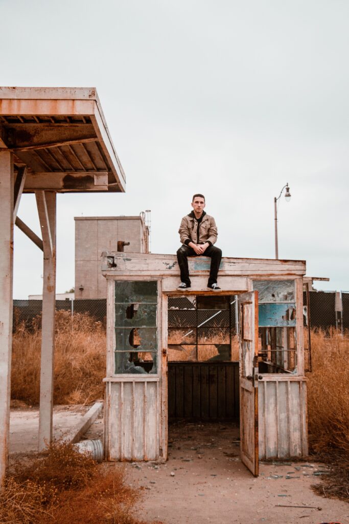 Person sitting on top of an abandoned weathered building surrounded by tall dry grass.