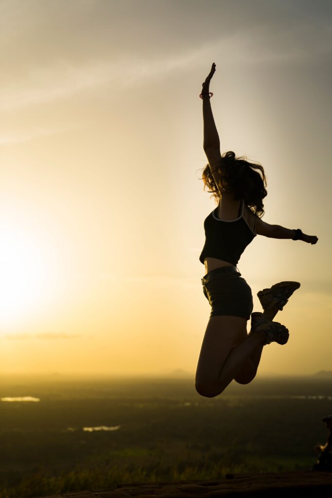 A silhouetted woman jumps into the air at sunset on a hilltop with her arms raised in celebration.