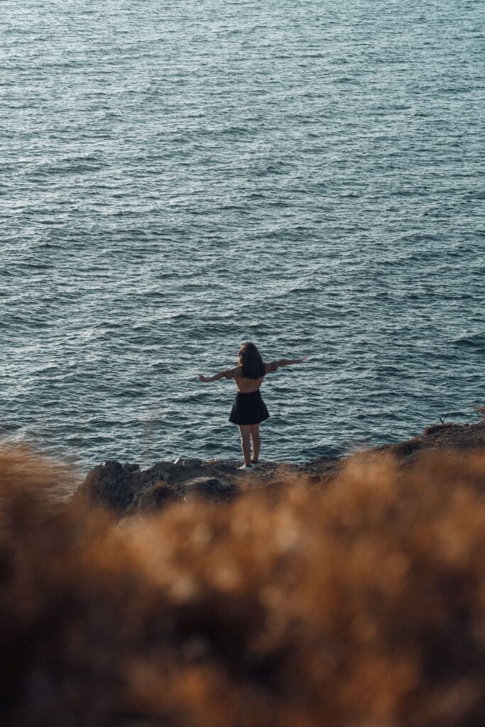 A woman stands on a rocky shoreline with her arms open toward the ocean, embracing the peaceful view.