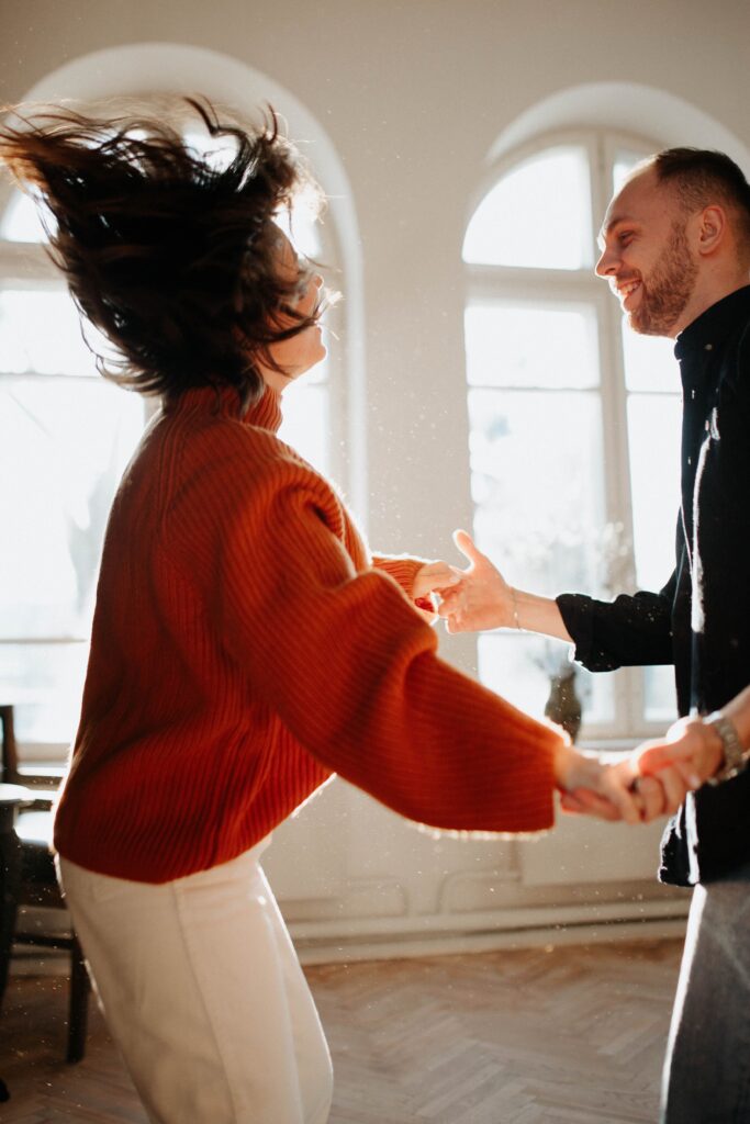 Two people smile and dance together in a sunlit room filled with warm afternoon light.
