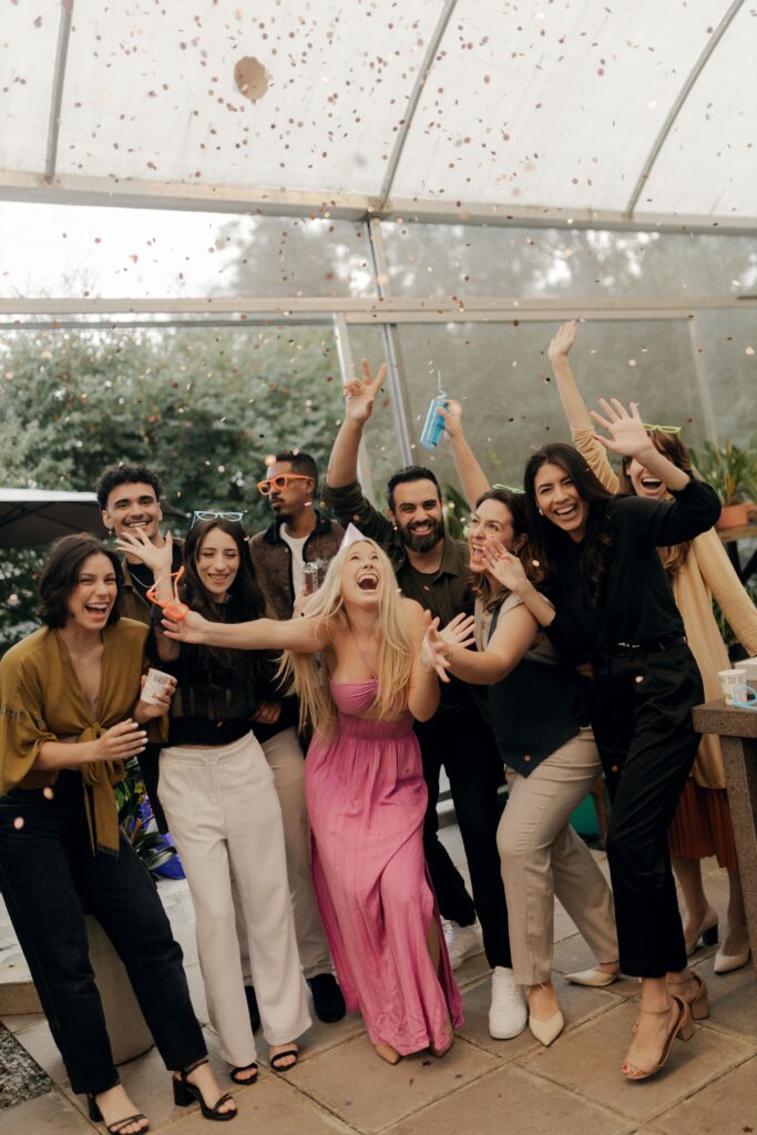A group of friends celebrates together as confetti falls around them, smiling, laughing, and posing for a joyful photo