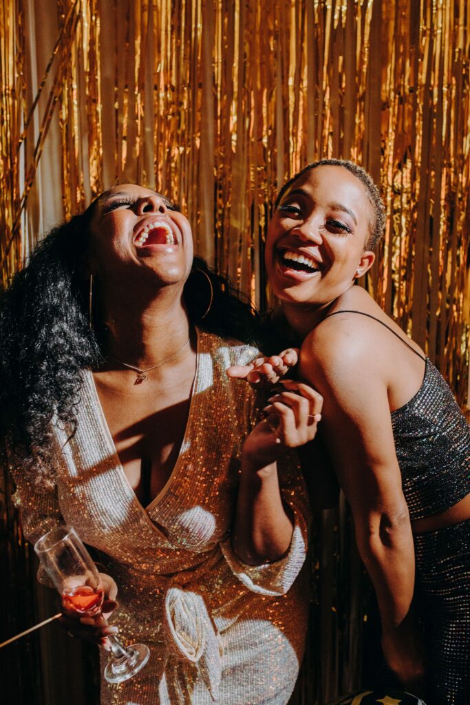 Two friends laugh together in sparkly outfits in front of shimmering gold streamers during a joyful celebration