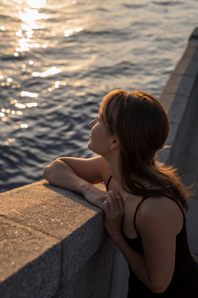 A young woman leans against a stone railing overlooking shimmering water at golden hour, smiling quietly as the sunset reflects across the waves.