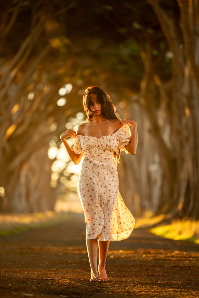 Woman in a floral dress walking barefoot beneath towering trees at sunset, creating a magical storybook atmosphere.