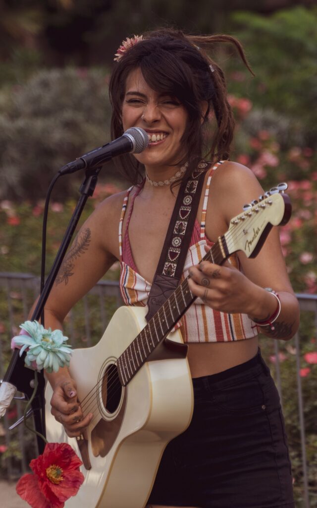 Smiling musician performing outdoors with an acoustic guitar in a flower-filled garden during golden hour.