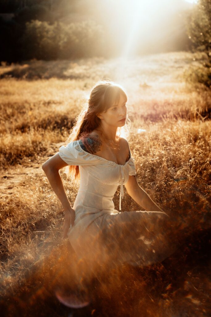Woman in a flowing white dress sitting in a golden field at sunset, creating a dreamy cinematic atmosphere that symbolizes dreaming big and romanticizing everyday life.